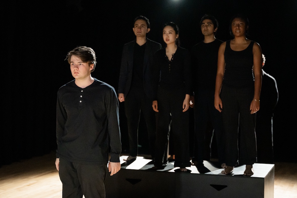 An actor stands on stage while a group of actors stand on a black riser behind him.